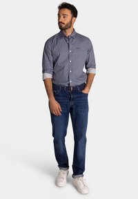 Blauwe button-up shirt met een klein gestippeld patroon, opgerolde mouwen; gecombineerd met donkerblauwe jeans en witte sneakers.