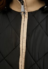 Sort quiltet jakke med en cremefarvet lynlås og krave. Tekstureret stof med et indviklet diamantmønster og kontrasterende kant.