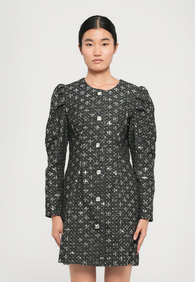 Woman wearing a fitted black sequin dress with long puff sleeves and large square buttons down the front, standing against a plain background.