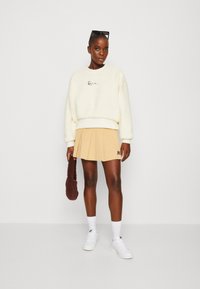 Felpa in pile crema con logo nero, mini gonna plissettata beige, sneakers bianche e borsa in pelle marrone. Occhiali da sole sulla testa.