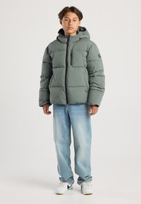 Giovane che indossa un giubbotto puffy verde con cappuccio, jeans azzurri chiari e scarpe bianche, di fronte a uno sfondo neutro.