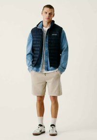 Chaleco acolchado azul marino sobre una chaqueta de mezclilla, combinado con una camiseta blanca y pantalones cortos beige. Zapatillas blancas con detalles negros, de pie sobre un fondo neutro.