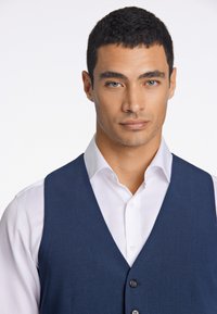 Marineblaue Weste über einem weißen Hemd; mit einem tiefen V-Ausschnitt, drei Knöpfen, glatter Textur und tailliertem Schnitt.