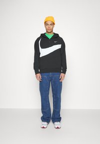 Čierna mikina s bielym dizajnom swoosh, zelené tričko pod ňou, modré džínsy a biele tenisky s červenými akcentmi; model nosí žltú čiapku.