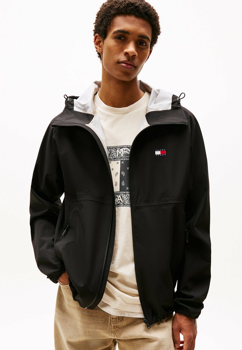 Giacca leggera nera con cappuccio grigio, dotata di un logo sul petto, polsini elasticizzati e chiusura con zip. Indossata sopra una leggera t-shirt grafica.