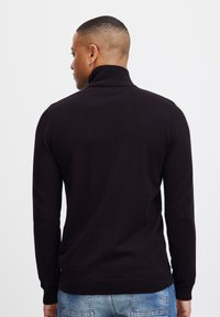 Suéter de punto negro con un cuello alto, mangas largas y puños acanalados. Textura suave, diseño ajustado y aberturas en los laterales.