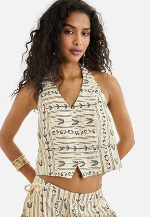 Mujer con cabello largo y rizado lleva un top halter beige y pantalones a juego con patrones en espiral y flechas negras, accesorios con una pulsera de oro.