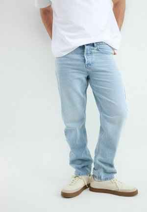 Osoba ubrana w jasnoniebieskie, luźne dżinsy, białe trampki z brązowymi podeszwami oraz białą koszulkę, stojąca na jednolitym tle.