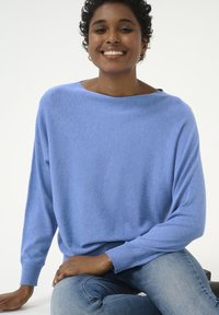 Lichtblauwe, losse trui met een brede halslijn, lange mouwen en geribbelde manchetten, gecombineerd met blauwe jeans. Zachte textuur, casual ontwerp.