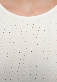 Λευκό τοπ με διαμάντι μοτίβο και μικρές καφέ κουκκίδες, στρογγυλή λαιμόκοψη και υφασμάτινη υφή, υπογραμμίζοντας περίτεχνη λεπτομέρεια.