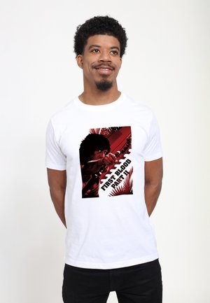 Hvid bomulds t-shirt med et grafisk print af en figur med en pil og teksten "FIRST BLOOD PART II" i sort og rød, omkranset af stiliserede palmeblade.