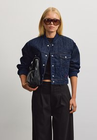 Donna con giacca di jeans scura oversize e occhiali da sole, che tiene una borsa di pelle nera su sfondo semplice.