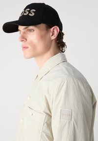 Black baseball cap with "BOSS" logo in grey; cream-colored shirt with visible buttons and a patch on the sleeve.