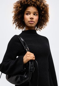 Γυναίκα με κατσαρά μαλλιά που φοράει μαύρο πλεκτό πουλόβερ, κρατώντας μια μαύρη δερμάτινη τσάντα στον ώμο της, κοιτάζοντας ελαφρώς προς το πλάι.