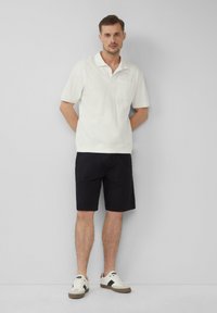 Camisa de cuello blanco de manga corta con rayas verticales, bolsillo en el lado izquierdo del pecho, combinada con pantalones cortos de color azul marino oscuro, usada con zapatillas blancas.