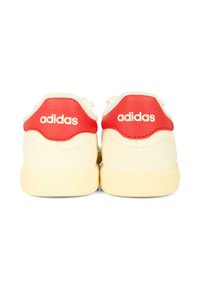 Baskets Adidas blanches et rouges, avec une tige en cuir, un dos texturé et des semelles en caoutchouc plates. Logo affiché sur le talon dans une couleur contrastante.