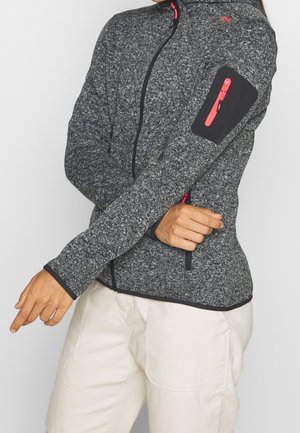 Personne portant une veste zippée grise avec des détails de fermeture éclair rouges sur la poche du bras et la poche latérale, associée à un pantalon beige clair.