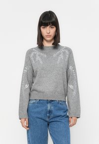Grå strikket genser med broderte bladmønstre på skuldrene, ribbet nederkant og lange ermer, kombinert med blå denimjeans.