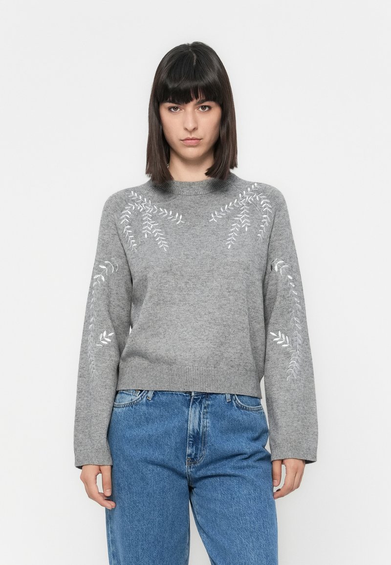 Grå strikket genser med broderte bladmønstre på skuldrene, ribbet nederkant og lange ermer, kombinert med blå denimjeans.