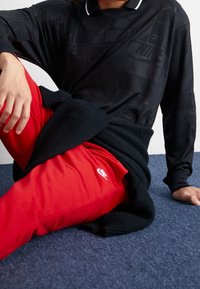 Person som sitter på en blå matta iförd en svart Nike Air långärmad tröja och röda Nike sweats, med en svart jacka knuten runt midjan.