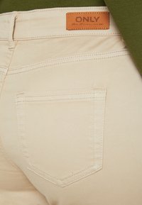 Beige jeans s sprednim zadnjim žepom, vidnim šivanjem in rjavim usnjenim oznako z imenom blagovne znamke "ONLY." Gladka tekstura.