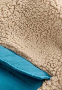 Tejido de piel sintética en color beige con una superficie texturizada y esponjosa, combinado con un material suave de color turquesa visible debajo.