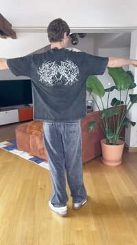 Asmuo stovintis viduje su išskėstomis rankomis, vilkintis juodą marškinėlį su grafika, pilkas škotų audinio kelnes, baltus sportbačius, šalia augalo ir sofos.