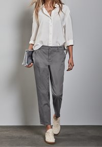 Grå lätta byxor med rak passform, parat med en vit knapplös skjorta och ljusa sneakers, hållande en silverfärgad kuvertväska.