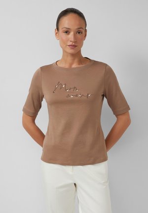 Kobieta z upiętymi włosami, ubrana w brązową bluzkę z krótkim rękawem z napisem "Mon cœur" oraz białe spodnie, stojąca na szarym tle.