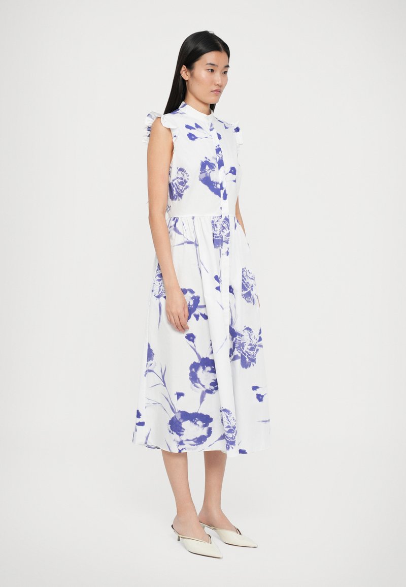 Floral print dress in white cotton with blue flowers, sleeveless design, ruffled shoulder accents, and midi-length. Paired with white shoes.