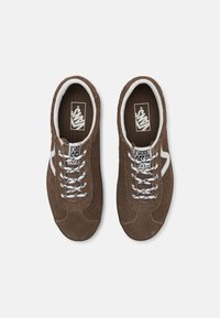 Ein Paar braune Wildleder-Vans-Sneaker mit weißen Seitenstreifen, weißen und schwarzen gemusterten Schnürsenkeln sowie mit Marken-Insocks auf einem weißen Hintergrund.