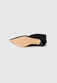 Scarpa nera in suede a punta con una superficie liscia, dettagli in elastico e una suola in gomma beige con un motivo testurizzato.