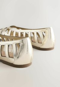 Chaussures plates dorées métalliques avec brides découpées et talons fermés, semelles marron sur fond blanc.