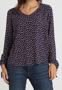 Donna che indossa una blusa a maniche lunghe blu scuro con piccoli motivi di foglie rosa, abbinata a jeans in denim blu.