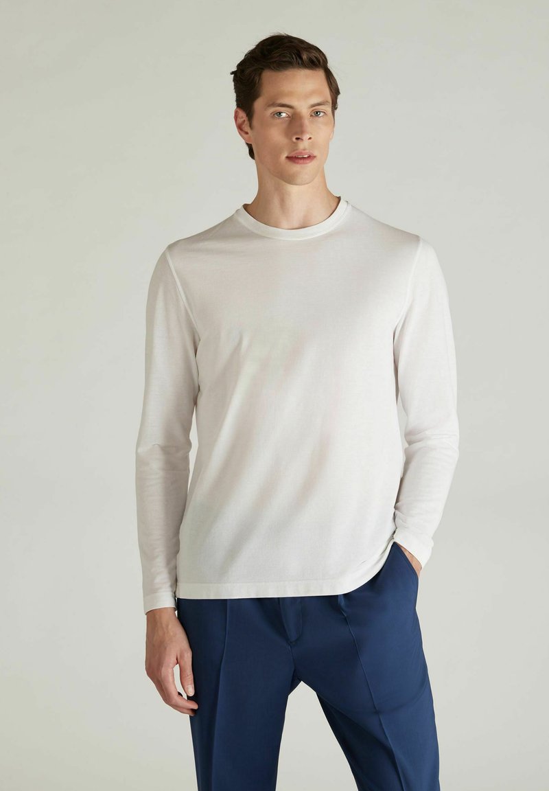 Junger Mann steht, trägt ein schlichtes weißes Langarmshirt und eine dunkelblaue Hose, eine Hand in der Tasche, vor einem hellgrauen Hintergrund.