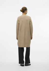 Dolg bež pleteni pulover s okroglo ovratnico in dolgimi rokavi, nosi se čez črne široke hlače, pogled od zadaj.