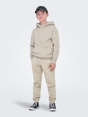 Garçon portant un sweat à capuche beige et un pantalon de survêtement, une casquette de baseball noire et des baskets noires avec des lacets blancs, debout les mains dans les poches.