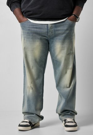 Person iført løstsiddende, falmede blå jeans med slidte pletter, sort-hvide sneakers og hænder i lommerne, stående mod en ensfarvet baggrund.