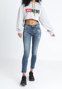 Sudadera gris recortada con un logo negro y rojo, combinada con jeans ajustados azules con un lavado desgastado y zapatillas deportivas negras.