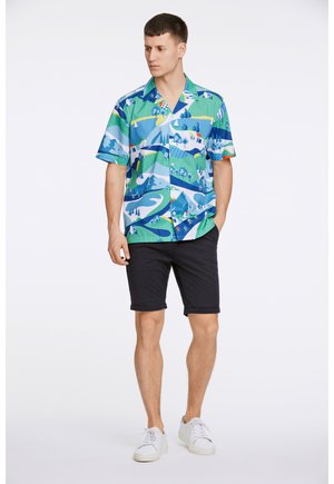 Camisa de botones de manga corta en azul y verde con un estampado de paisaje; combinada con pantalones cortos negros y zapatillas blancas.