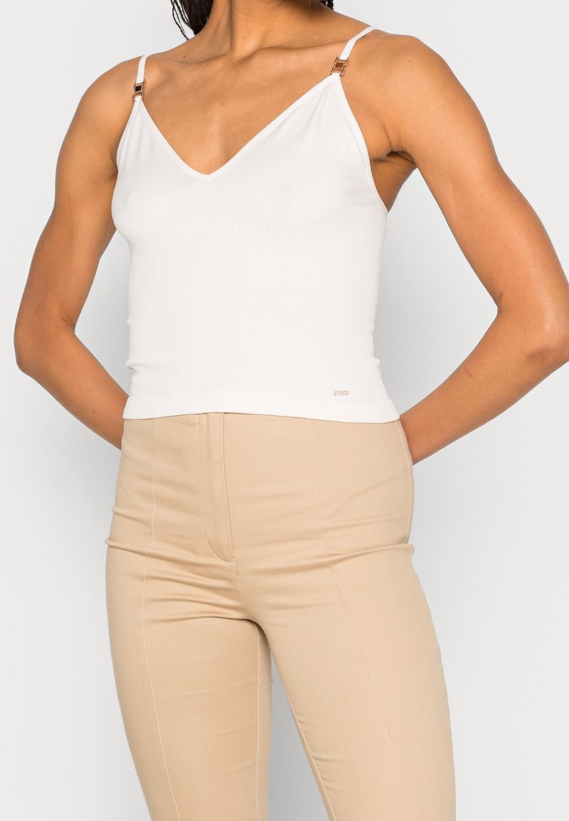 Top a maniche corte bianco con collare a V, spalline sottili regolabili con dettagli metallici dorati, abbinato a pantaloni beige a vita alta.