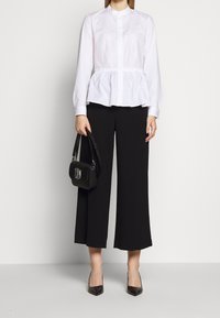 Donna che indossa una blusa peplo bianca a maniche lunghe, pantaloni neri a gamba larga e cropped, tacchi a punta neri e tiene in mano una borsa nera.