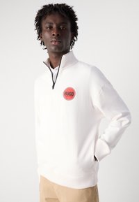 White zip-up sweatshirt with ribbed cuffs and collar, featuring a circular red "HUGO" logo on the chest. Smooth fabric texture.