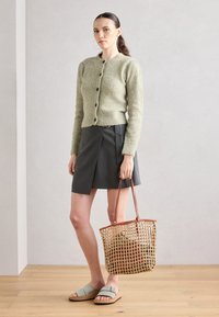 Grön stickad cardigan med knappar, grå omlottkjol, ljusgröna sandaler och vävd korgväska med läderhandtag. Släta texturer, modern design.