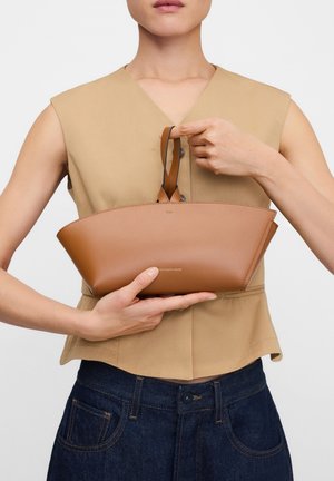 Femme portant un haut beige sans manches et un jean foncé, tenant un petit sac à main en cuir marron avec des poignées supérieures devant la poitrine.