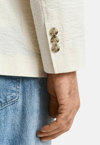 Blazer de couleur crème avec une finition texturée, doté de trois accents en métal argenté aux poignets et d'une coupe ajustée sur un denim.