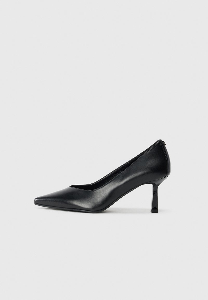 Chaussure à talon aiguilles noir à bout pointu, talon moyen, cuir lisse, côtés échancrés, sur un fond clair uni.