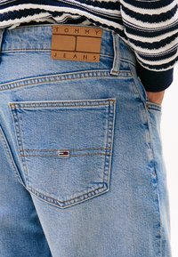 Heledad sinine denimpüksid, millel on pruun nahklabel "TOMMY JEANS" ja väike punase, valge ja sinise embleem tagakotis.
