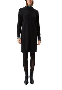 Schwarzes gestricktes Kleid mit hohem Kragen, langen Ärmeln, gerippten Bündchen und gerader Silhouette. Kombiniert mit schwarzen Stiefeletten.
