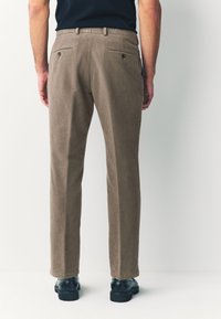Pantalon marron clair à texture lisse, avec deux poches arrière et une coupe droite. Porté avec des chaussures noires pour le contraste.